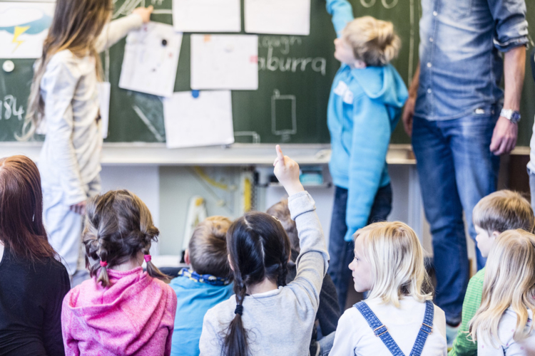 Kinder sitzen vor einer Tafel und melden sich