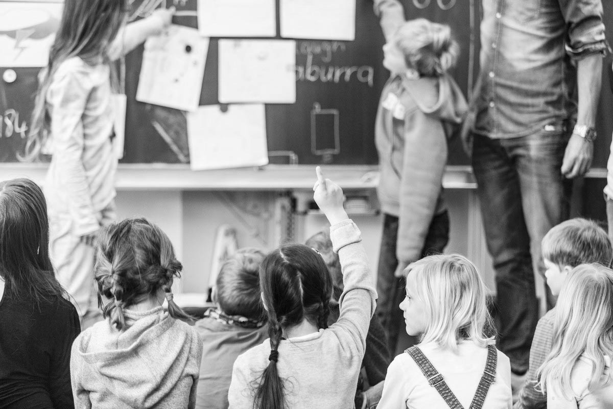 Kinder sitzen vor einer Tafel und melden sich